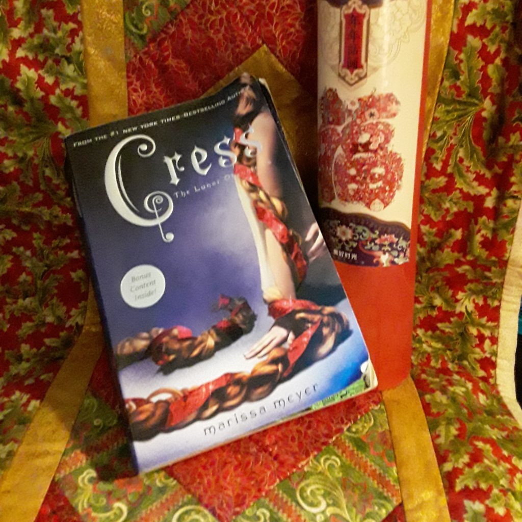 A paperback copy of Cress sits on a Christmas quilt next to a red document tubedecorated for Chinese New Year. There's lots of red.