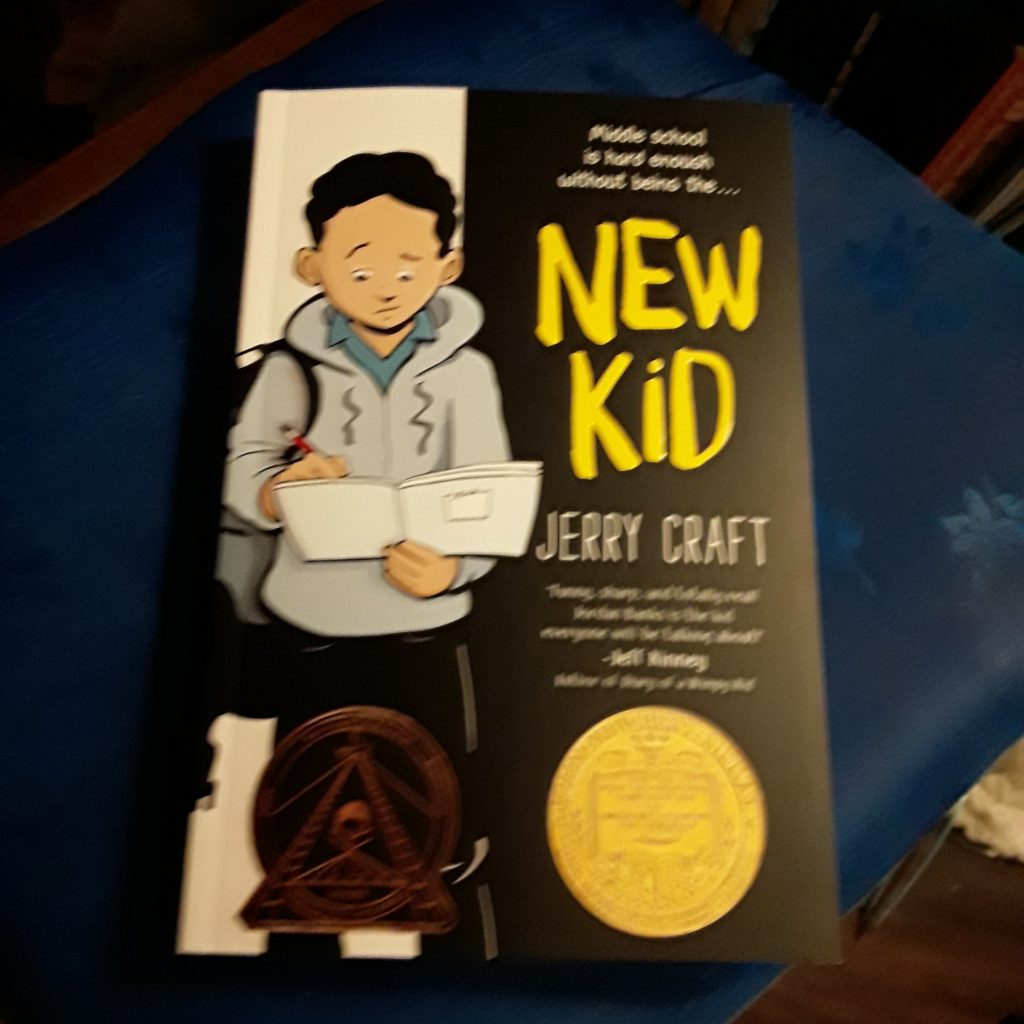 New Kid--by Jerry Craft The book sits on a chair On the cover, a kid in a gray hoodie and black slacks scribbles in a notebook