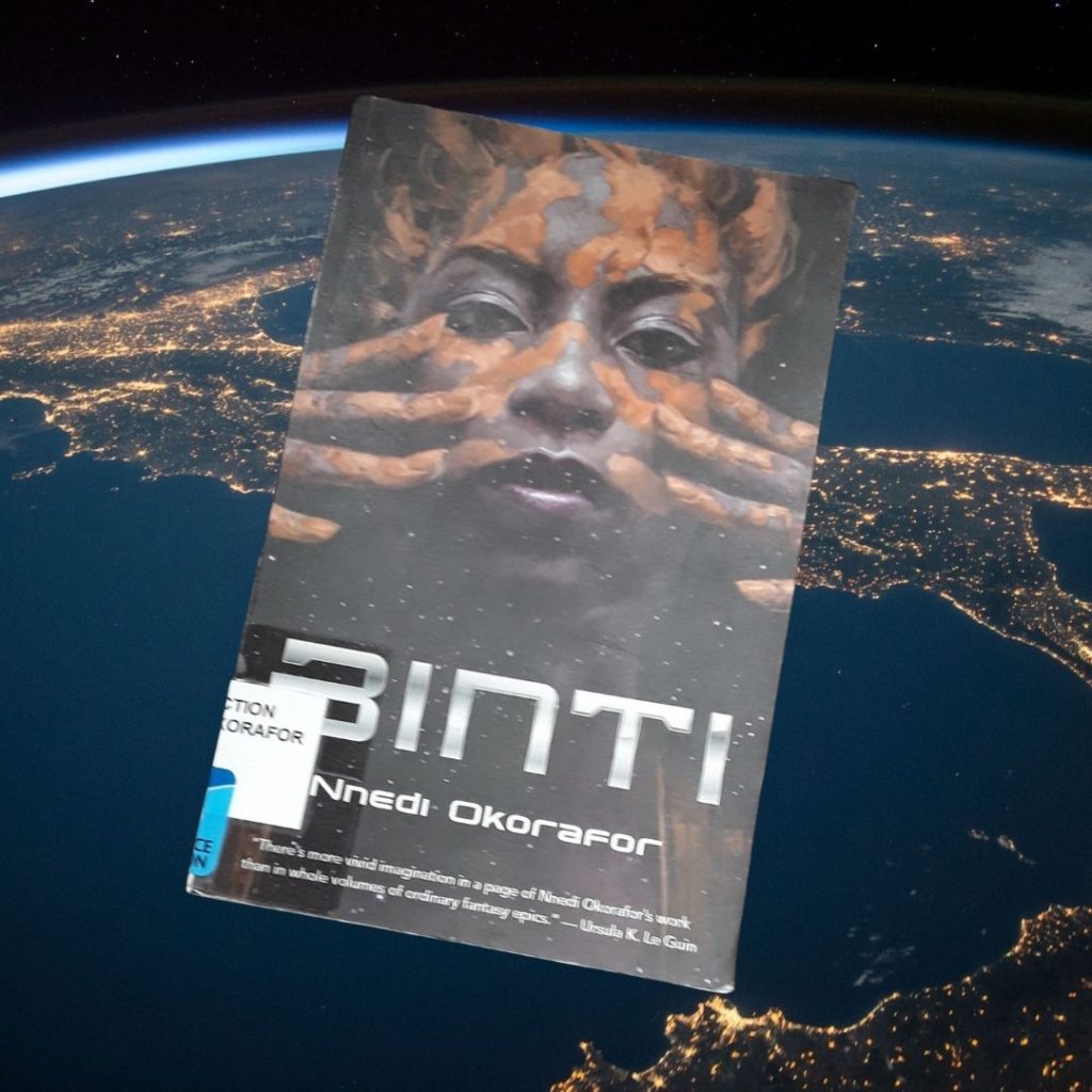 A library copy of Binti floats over Earth as if the book is in space. On the cover, a brown-skinned woman marks patterns in an orangish red on her face.