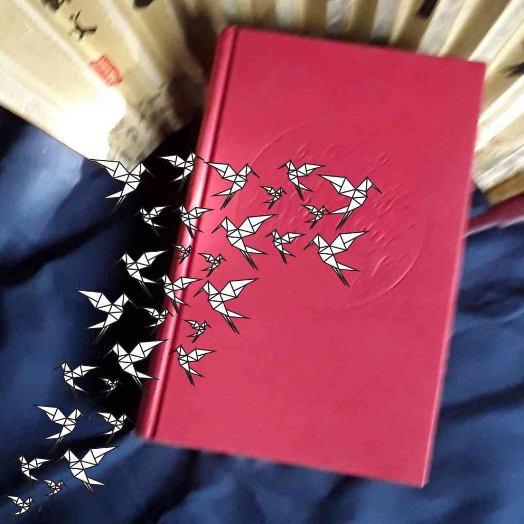 A hardback copy of The Girl who Drank the Moon--without its jacket-- sits atop a fan, and pictures of paper cranes swarm above it.