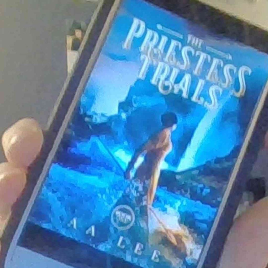 The Priestess Trials cover on a cell phone. A woman in a leather dress holding a spear looks out over a river toward a waterfall.