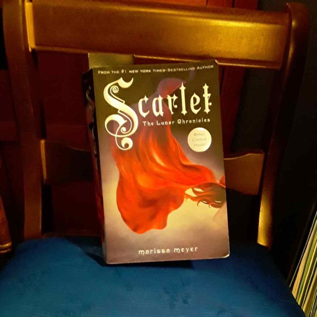 A paperback copy of Scarlet sits on a ladderback chair. On the cover, red fabric flows.